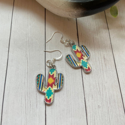 Colorful cactus-shaped earrings on a wooden surface with a plant in the background