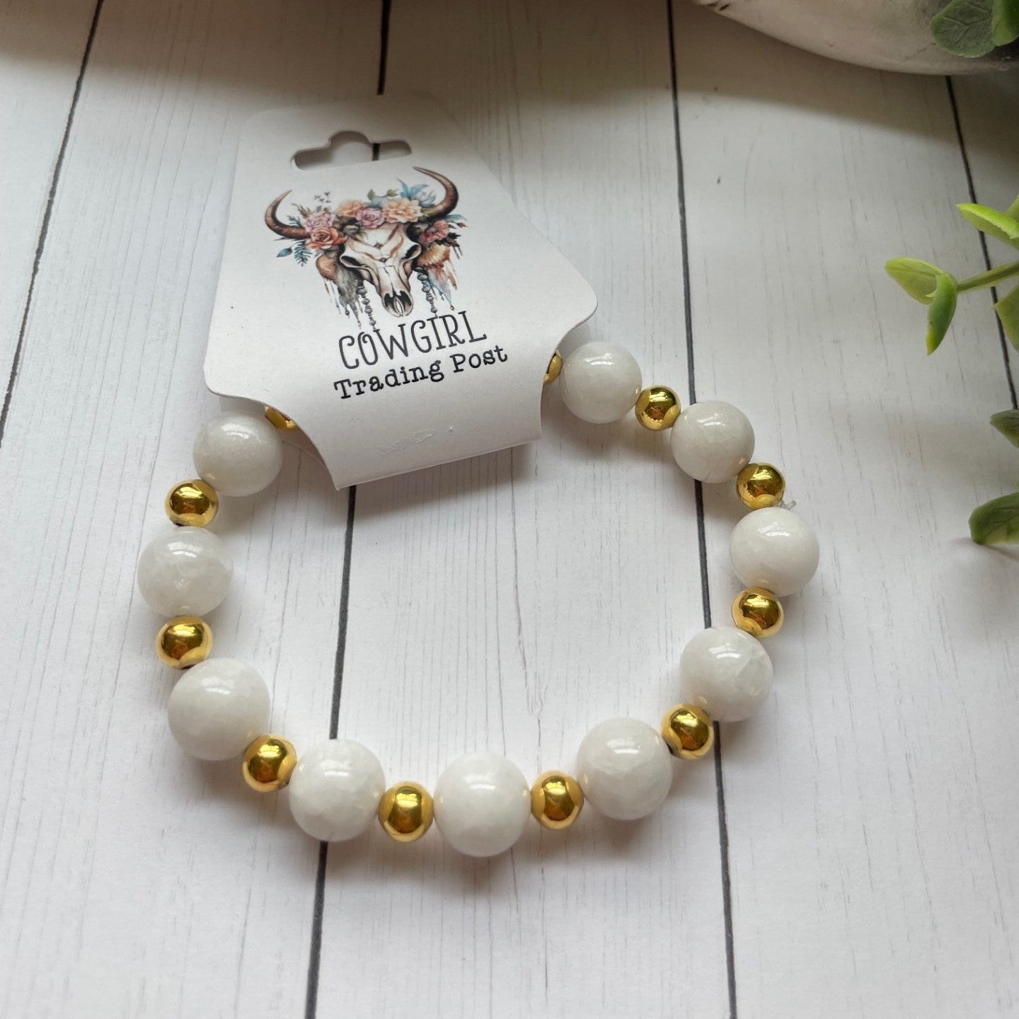 White beaded bracelet with gold accents on a white wooden surface, branded 'The Cowgirl Trading Post'.