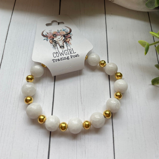 White beaded bracelet with gold accents on a white wooden surface, branded 'The Cowgirl Trading Post'.