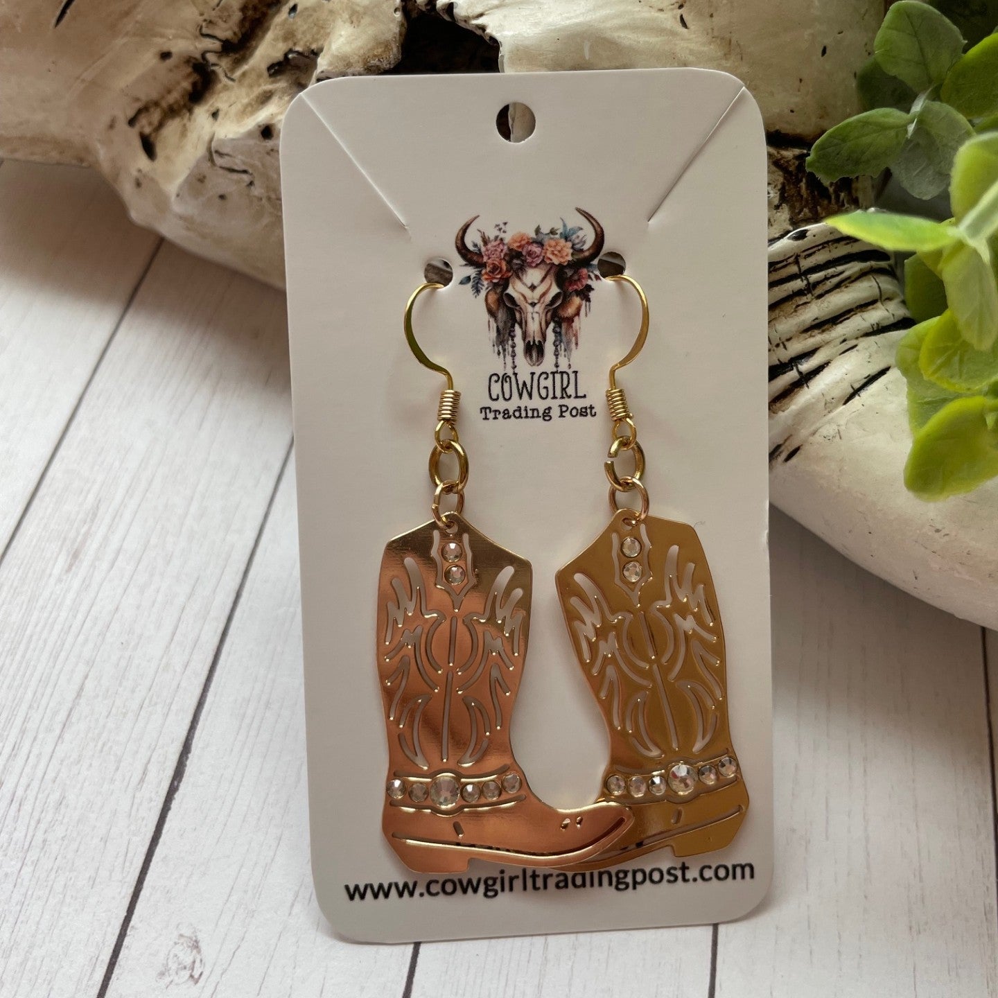 Cowboy boot earrings on a card with 'Cowgirl Trading Post' branding, placed on a wooden surface.