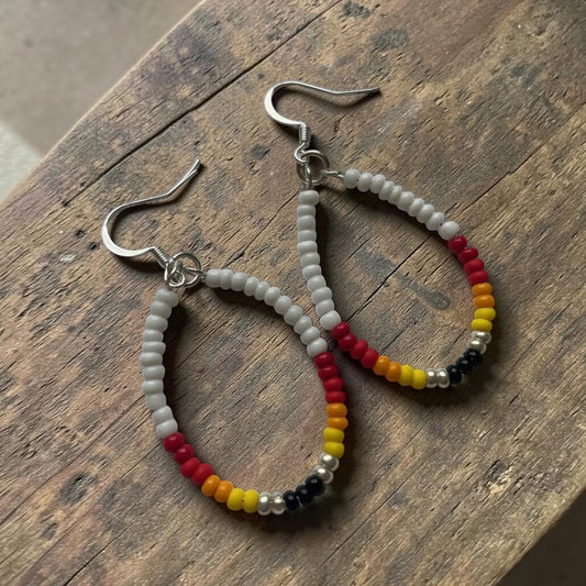 A pair of handmade hoop earrings with silver-toned fishhook wires, featuring white seed beads and a multicolor section of red, yellow, black, and silver beads, displayed on a rustic wooden surface.