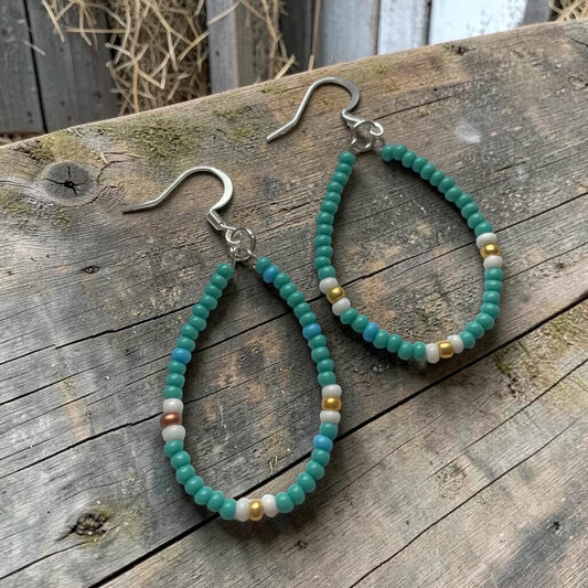 Teardrop-shaped beaded dangle earrings with turquoise-green beads, small white beads, and a few metallic gold and copper beads, hanging from silver fishhook wires on a weathered wooden surface.