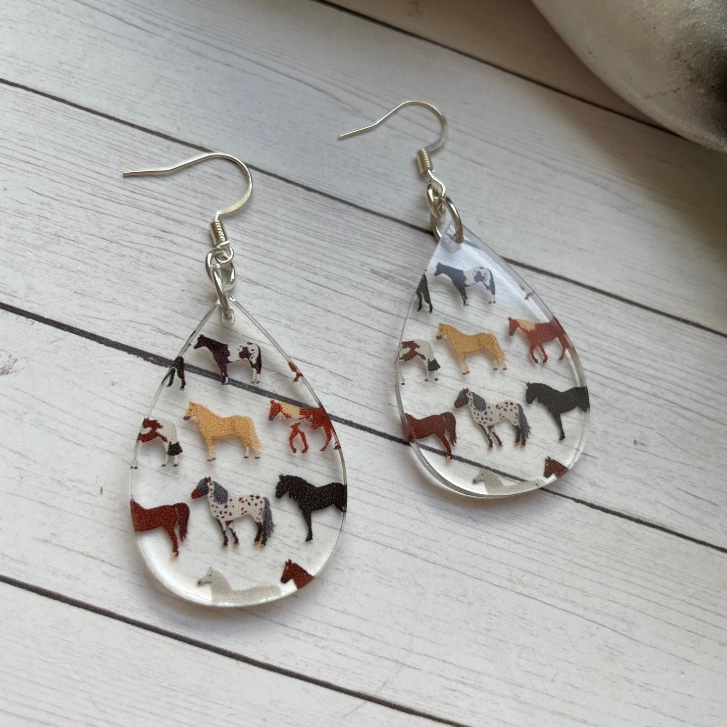 Teardrop-shaped earrings with horse illustrations on a wooden surface