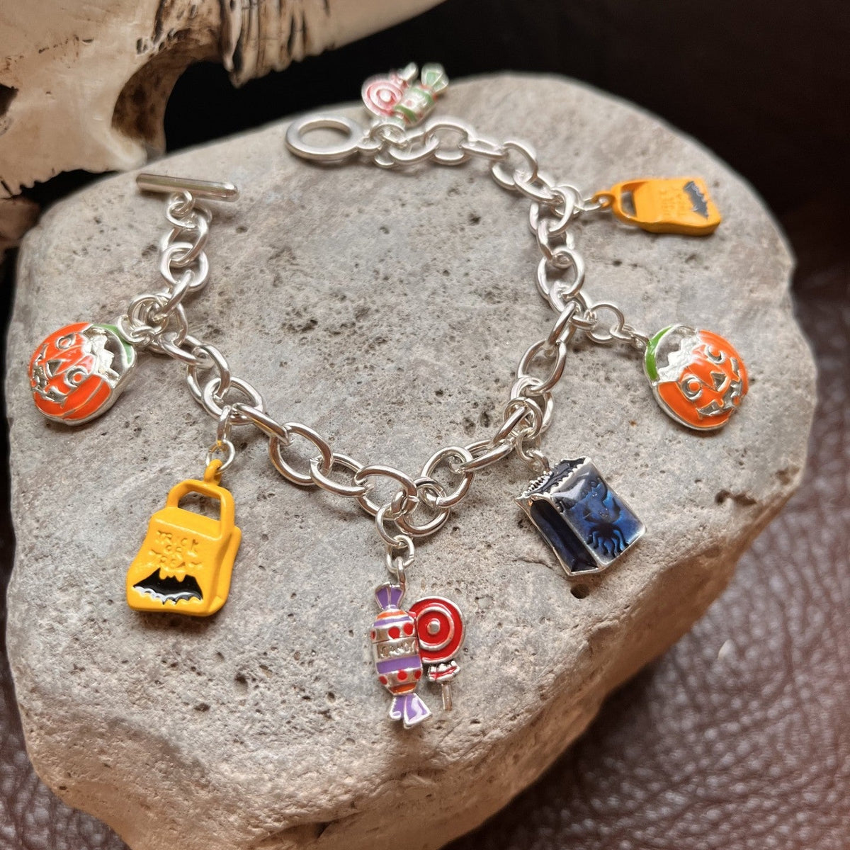 A silver charm bracelet displayed on a gray stone, featuring colorful enamel charms shaped like a jack-o'-lantern, a yellow car, a blue book, a candy cane, and a small orange item, with a white horn prop in the background.