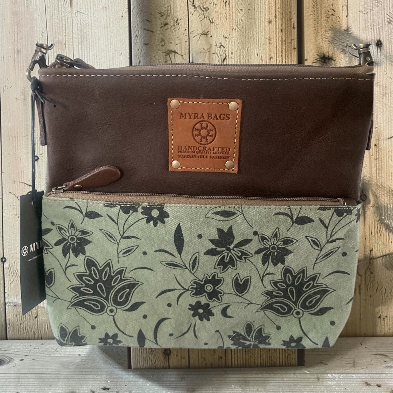 A Myra Bags crossbody bag with a dark brown leather top half and a sage green canvas bottom half featuring a black floral pattern. A tan leather patch on the front reads 'MYRA BAGS HANDCRAFTED'.
