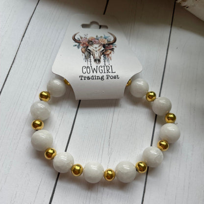 White beaded bracelet with gold accents on a white wooden surface, branded 'The Cowgirl Trading Post'.