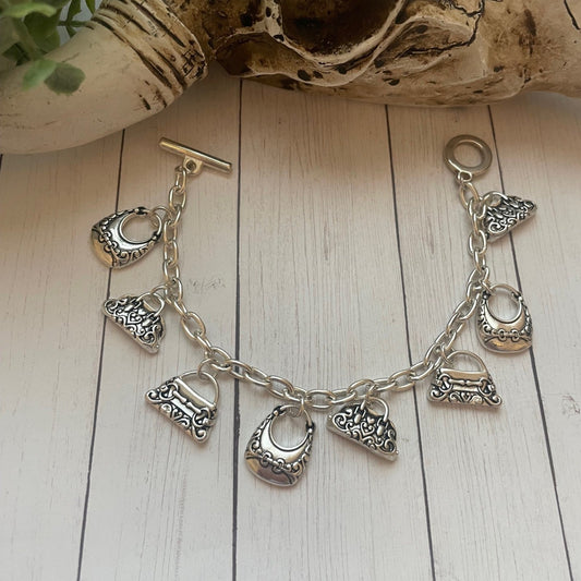 A silver-toned charm bracelet featuring multiple small handbag and purse-shaped charms, displayed on a white wood plank surface with decorative props including a skull and greenery in the background.