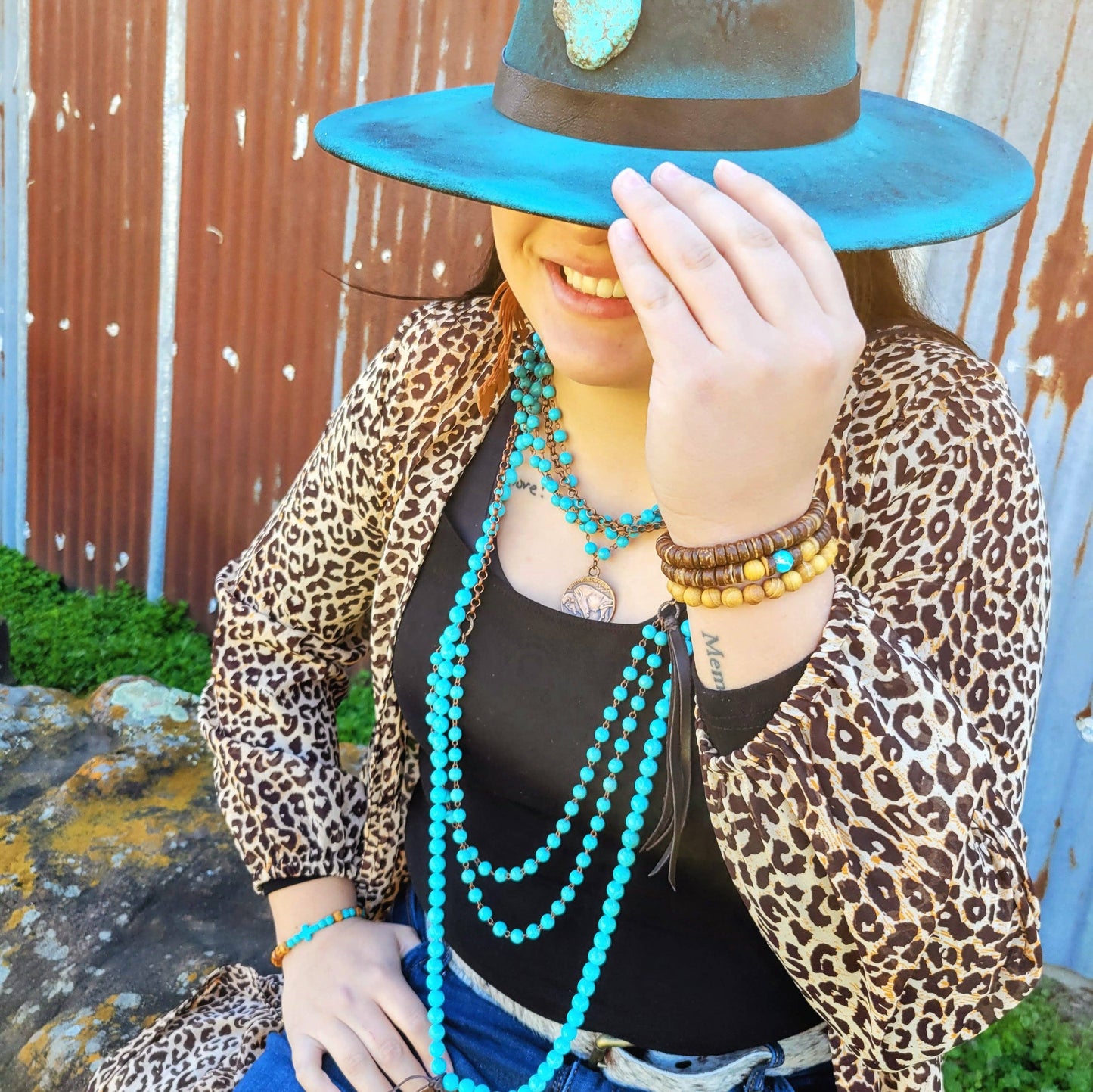 Person wearing a leopard print jacket and turquoise hat with multiple necklaces, sitting against a rustic wall.