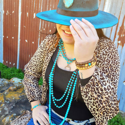 Person wearing a leopard print jacket and turquoise hat with multiple necklaces, sitting against a rustic wall.