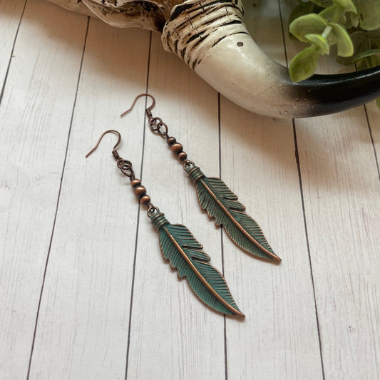 Feather-shaped earrings on a wooden surface with a plant in the background