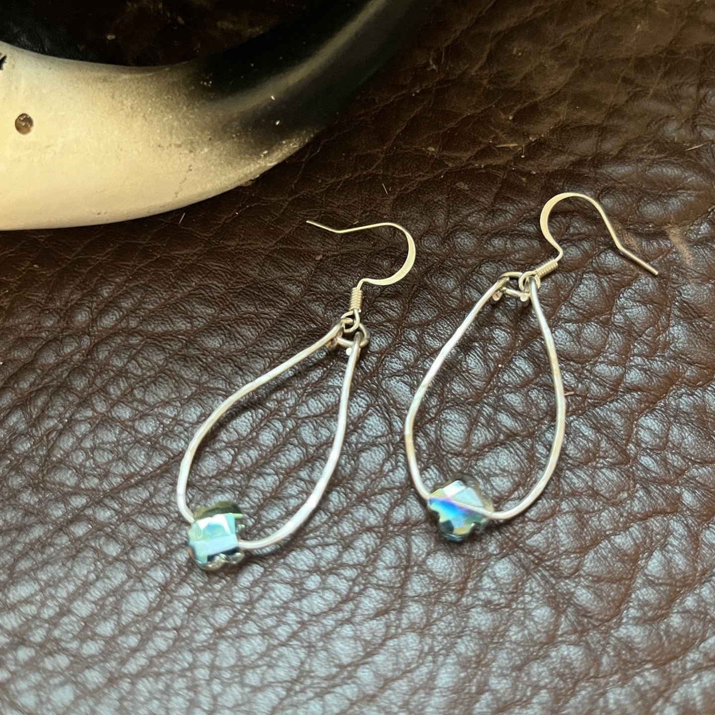A pair of silver teardrop dangle earrings with small iridescent blue-green square stones, displayed on a textured brown leather surface.