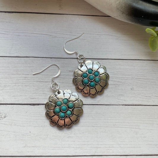 Floral earrings with turquoise centers on a wooden surface with a skull and plant in the background