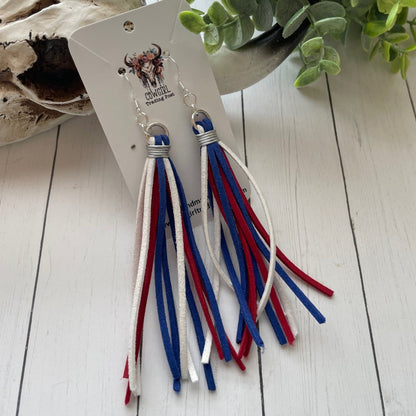 Tassel earrings with red, white, and blue strands on a card with branding, placed on a wooden surface with greenery.