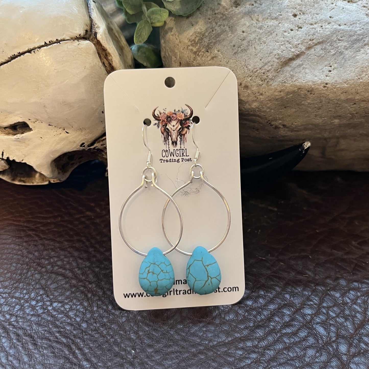 A pair of silver hoop earrings with turquoise teardrop charms, displayed on a white cowgirl-themed card. The card is placed on a dark brown textured surface with a skull and stone props in the background.