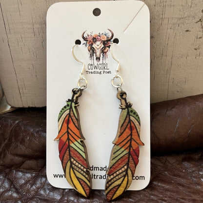 A pair of feather-shaped dangle earrings displayed on a white Cowgirl Trading Post card, resting on a brown leather surface against a rustic wood background.