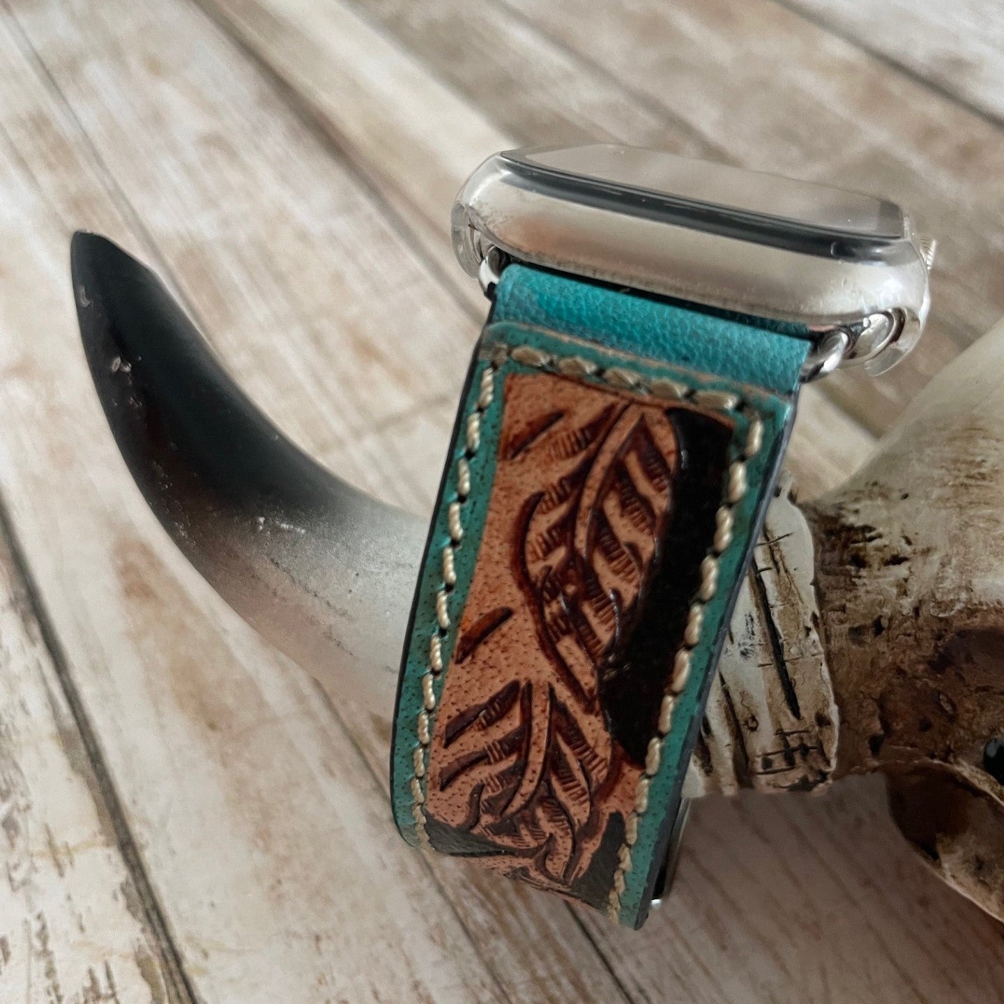 Watch with a turquoise leather strap featuring leaf patterns on a wooden surface.