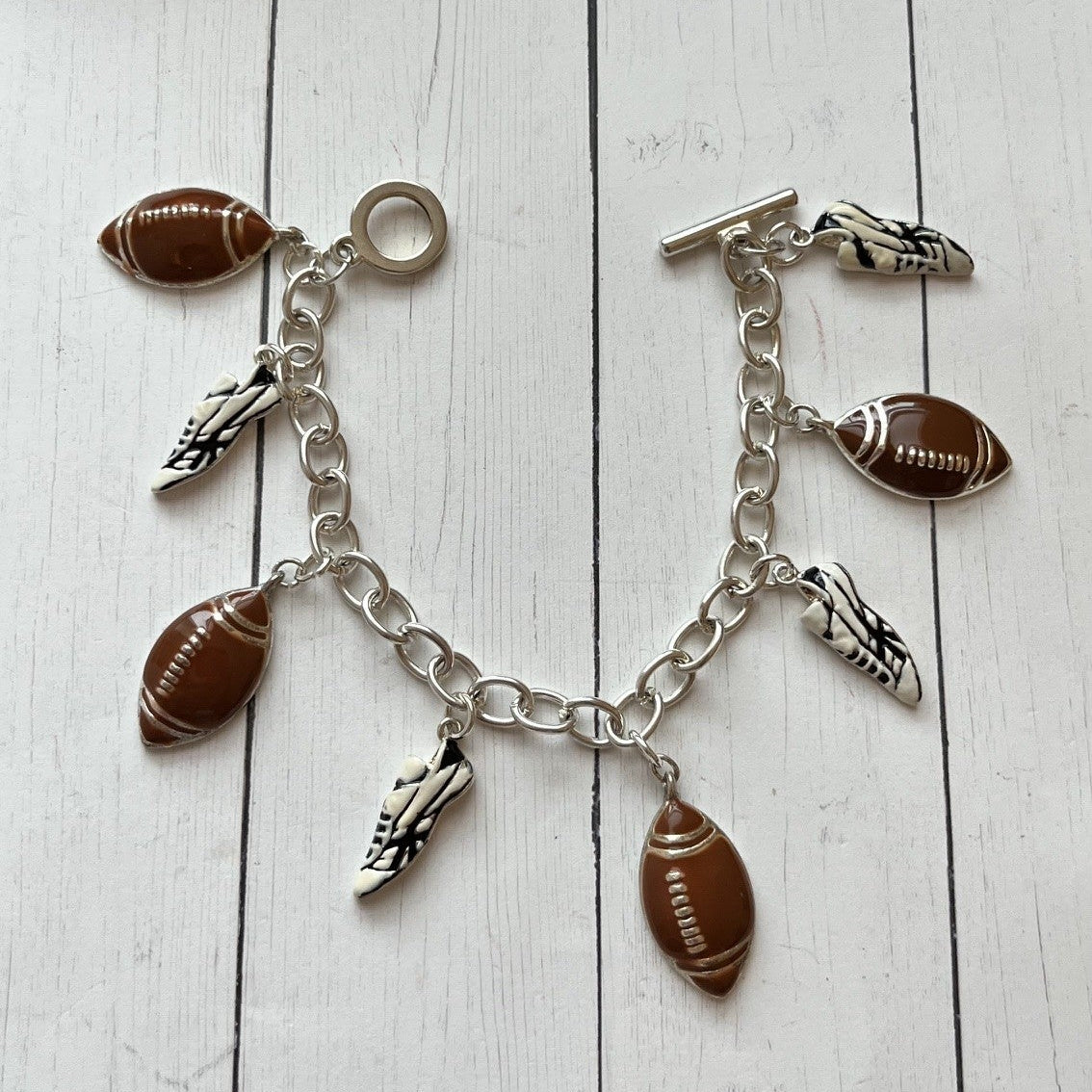 A silver-tone chain bracelet with football and shoe charms, displayed on a white wood plank surface.