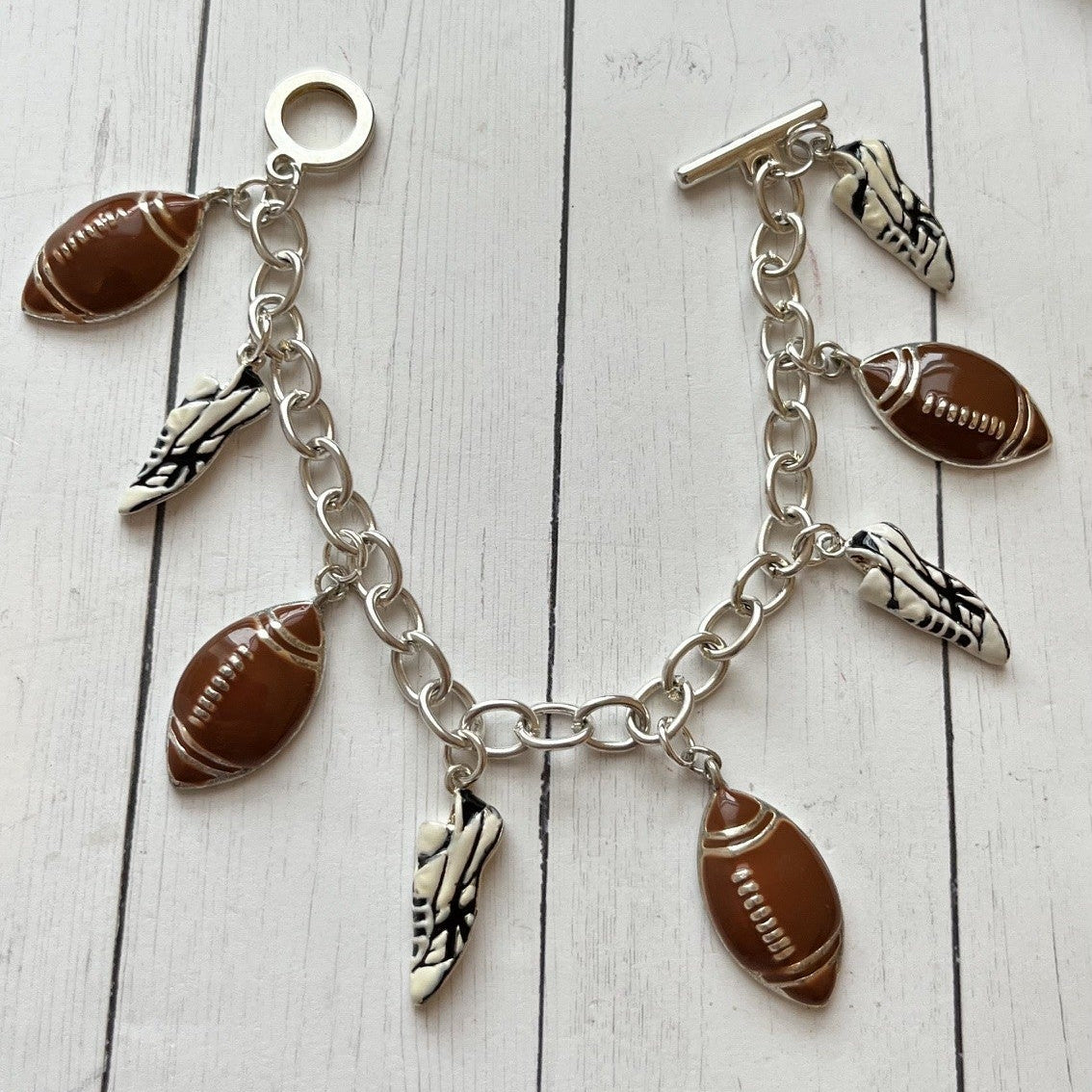 A silver-tone chain bracelet with football-shaped charms, feather charms, and a small silver ring, displayed on a white wood surface next to a dark brown leather item.