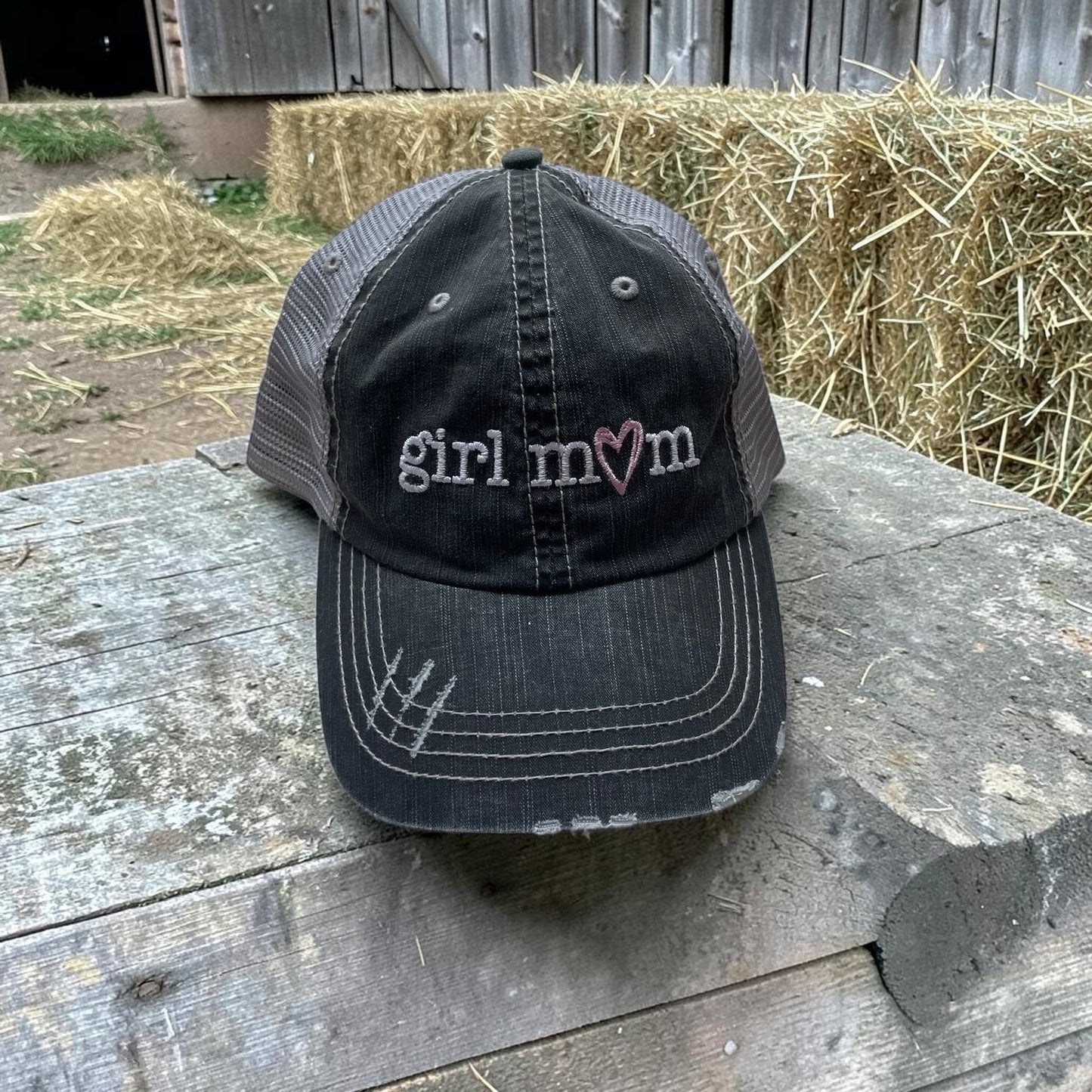 Black cap with 'girl mom' text on a wooden surface with hay bales in the background