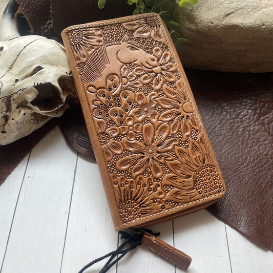 A brown leather zip-around wallet with embossed floral and bird motifs, styled on a white wood surface with a skull, dark leather, and stone props.