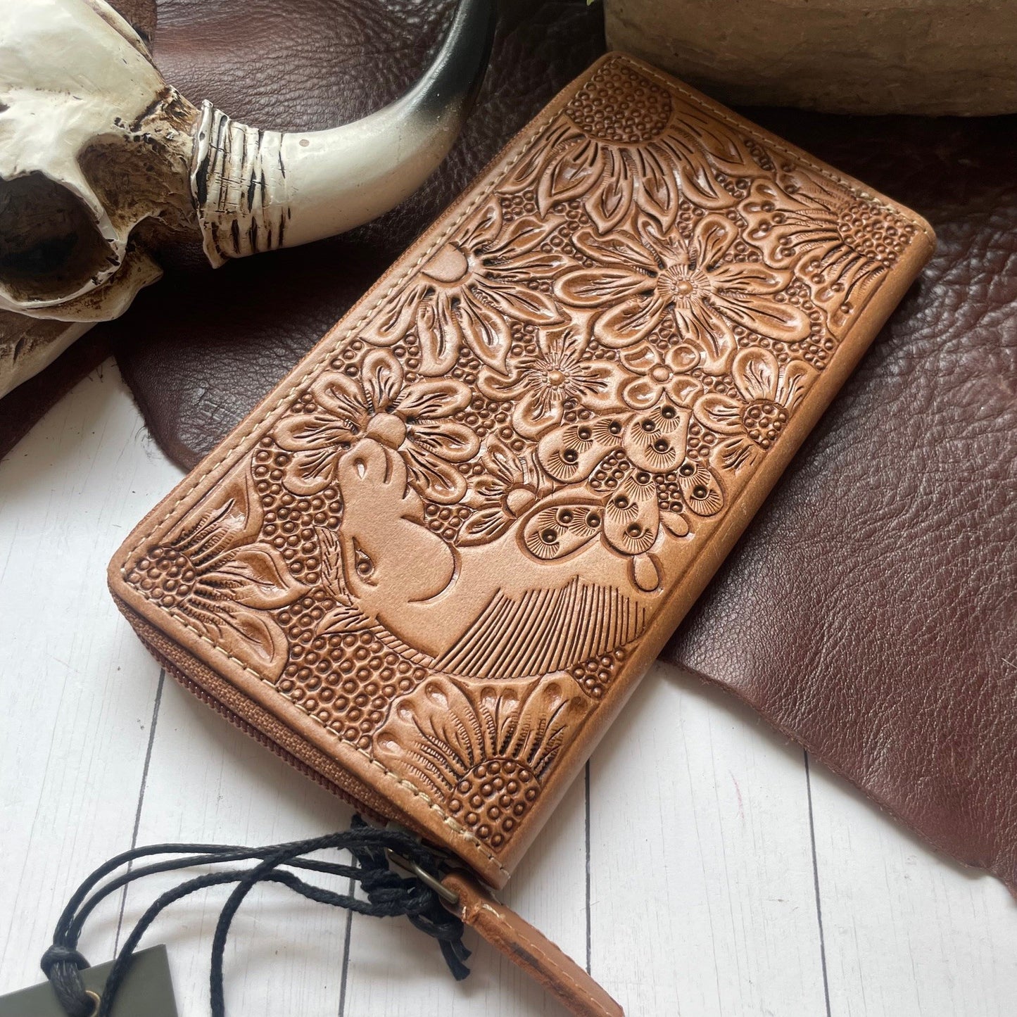 A tan leather zip-around wallet with embossed floral and bird designs, styled on a white wood surface with a brown leather pad, a skull prop, and a wooden object in the background.