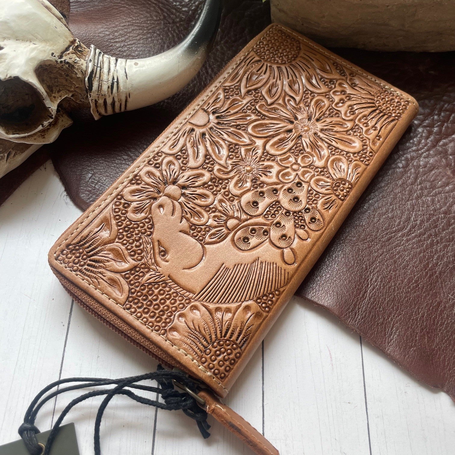 A tan leather zip-around wallet with embossed floral and bird designs, styled on a white wood surface with a brown leather pad, a skull prop, and a wooden object in the background.