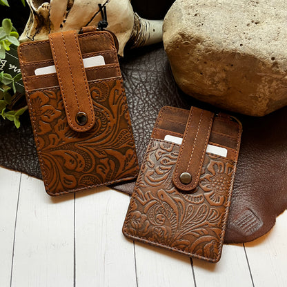 Two brown embossed leather card holder wallets with snap closures, displayed on a dark textured mat with rustic props including a rock, wood, and small plant.