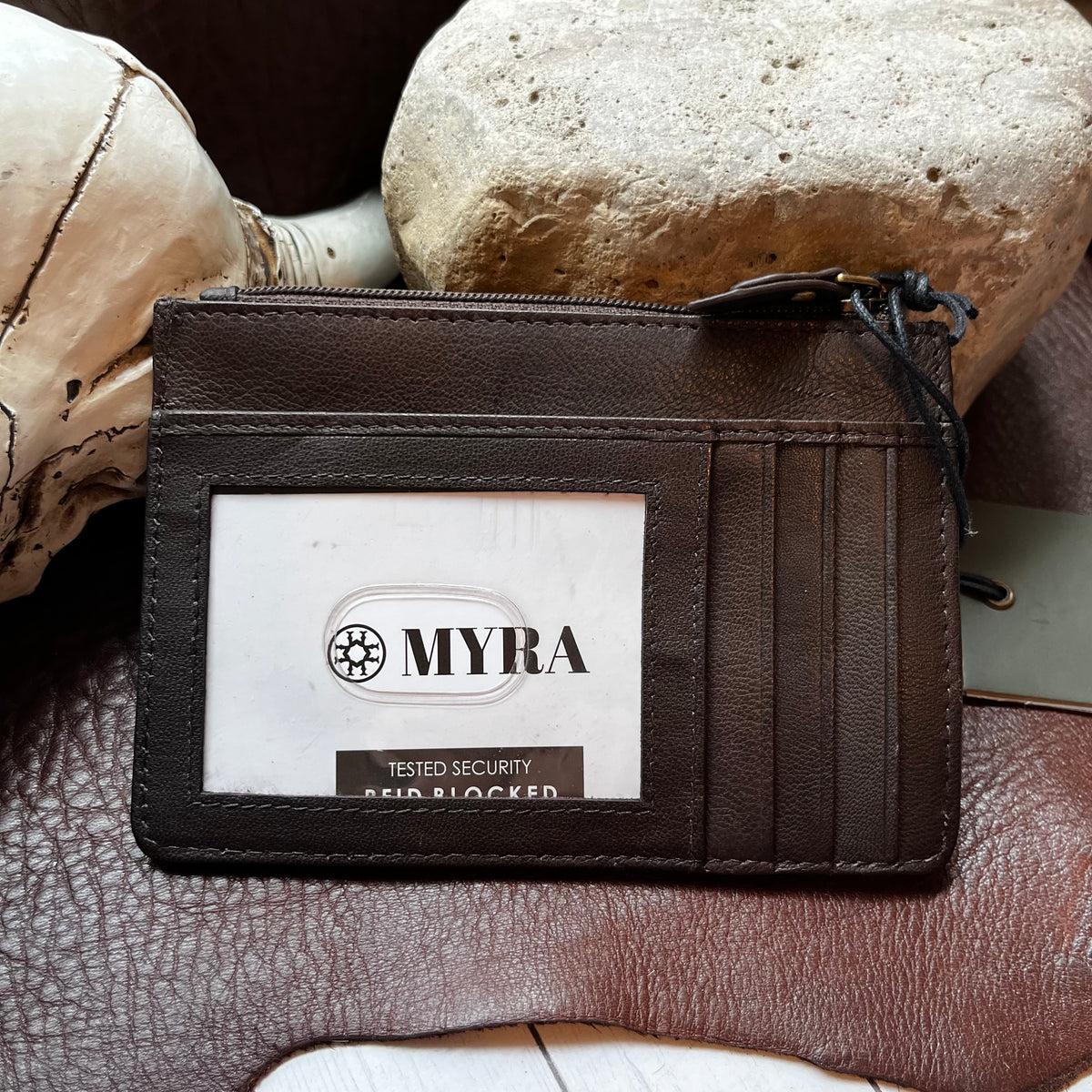 A dark brown leather card holder wallet with a clear ID window displaying a white 'MYRA' security badge, styled on a brown leather surface with a skull and stone props.