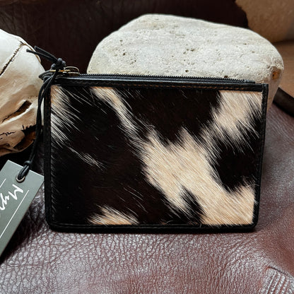 A small zip pouch with a black and beige cowhide pattern, resting on a brown leather surface with a stone prop behind it.