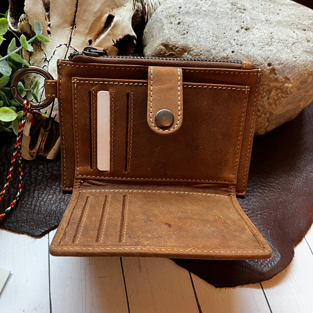 An open brown leather wallet with multiple card slots, a zipper compartment, and a snap-button closure, styled on a dark leather pad with a skull, rock, and greenery in the background.