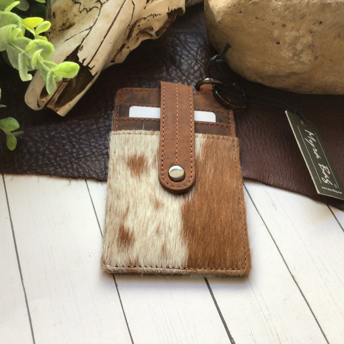 A small bifold card holder wallet made of brown and white cowhide with a tan leather strap and snap closure, displayed on a white wood surface with leather and decorative props in the background.