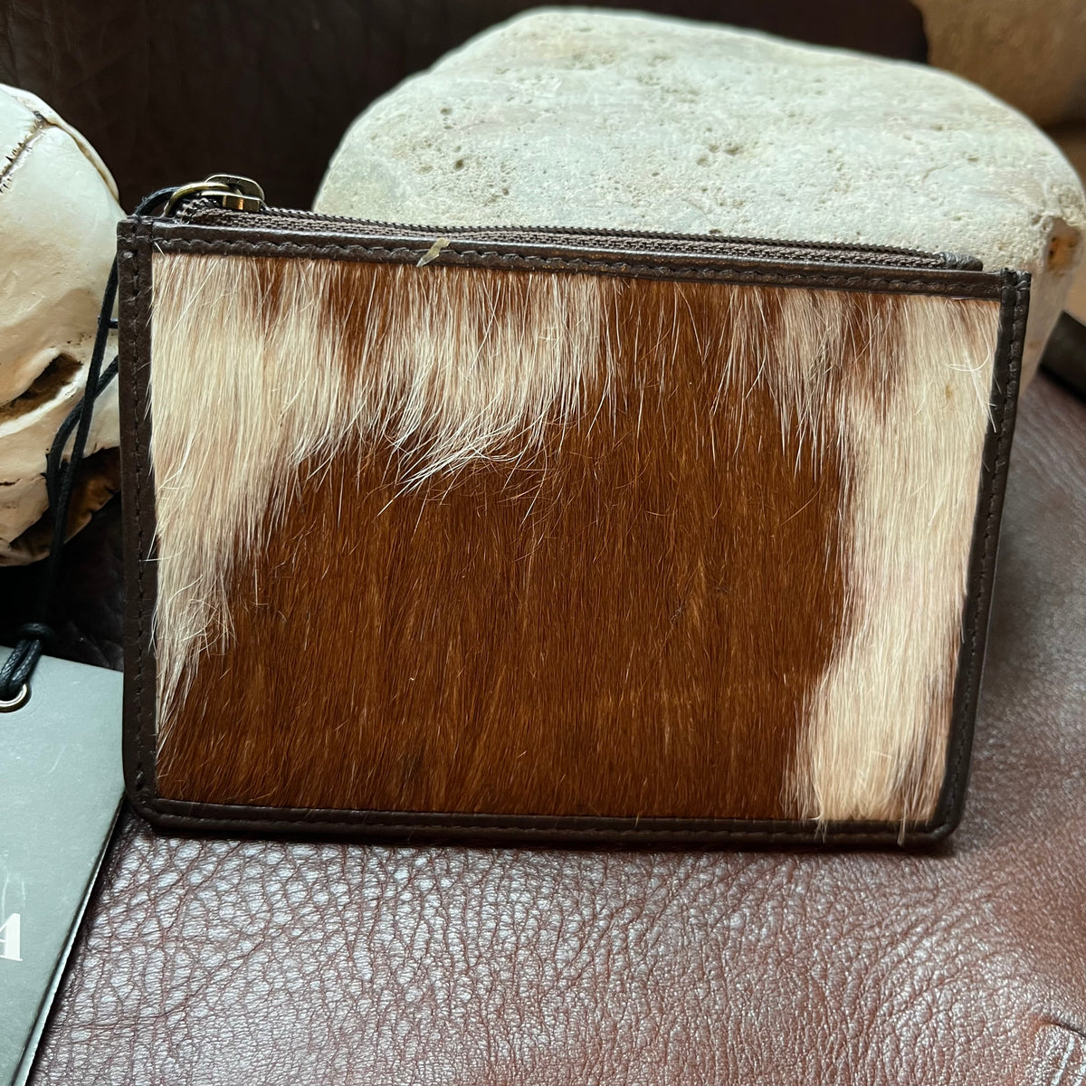 A small zip-around pouch or clutch made from brown and cream cowhide with dark brown leather trim, photographed on a brown textured surface with stones in the background.