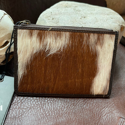 A small zip-around pouch or clutch made from brown and cream cowhide with dark brown leather trim, photographed on a brown textured surface with stones in the background.