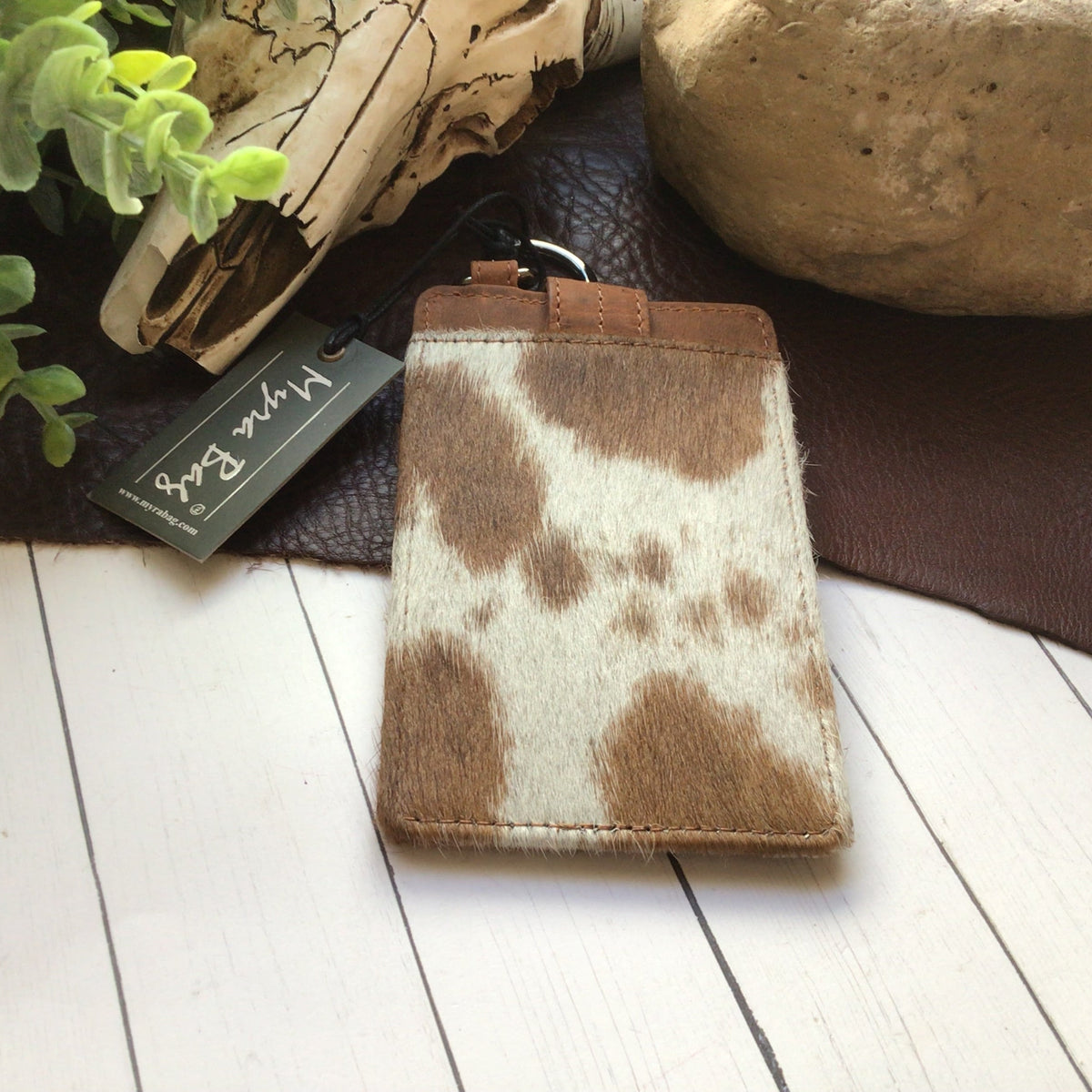 A small leather card holder or pouch with a brown and cream cowhide pattern, displayed on a white wood surface with a leather tag reading 'Wine Red' and rustic decor in the background.