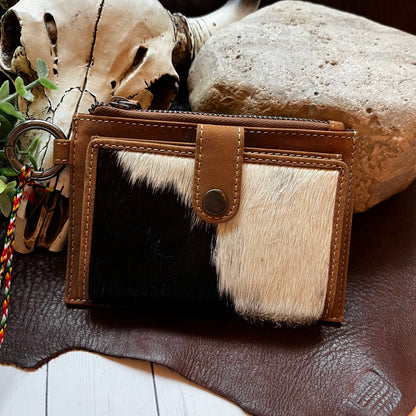 A brown leather and black-and-white cowhide wallet with a snap-button closure and attached key ring, styled on a leather surface with a skull, rock, and small plant in the background.