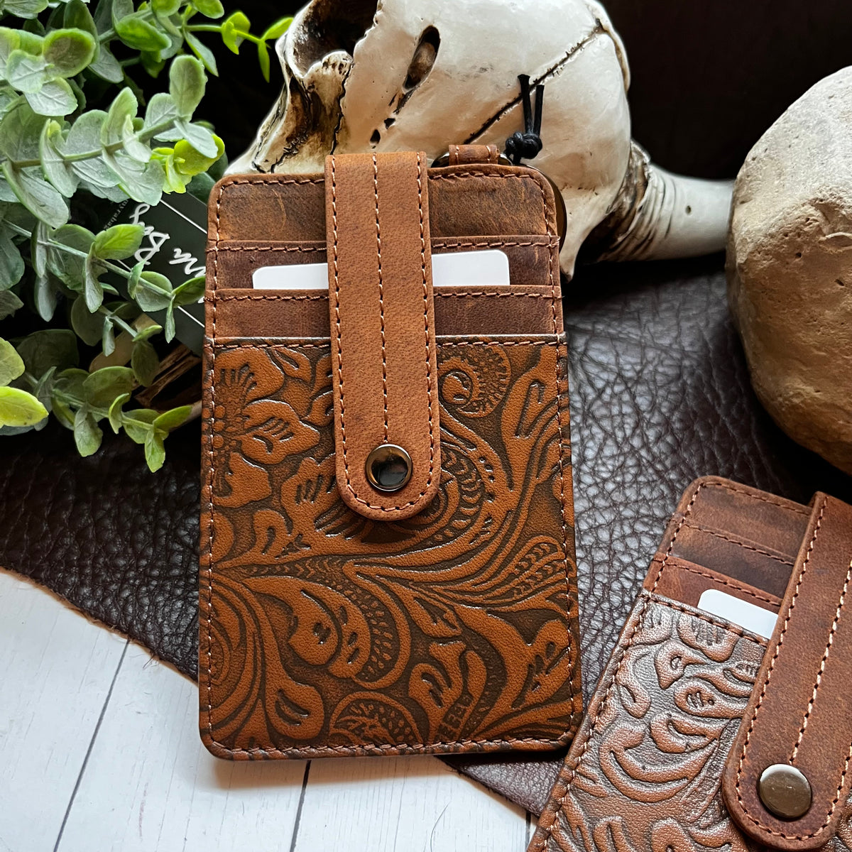A brown tooled leather card holder wallet with an embossed floral and scroll pattern, displayed on a dark leather mat with a skull, plant leaves, and a stone prop.