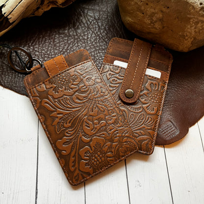 Two brown tooled leather card holders with floral embossing, one closed and one open to show card slots and snap closure, displayed on a white wood surface with leather and rock props.