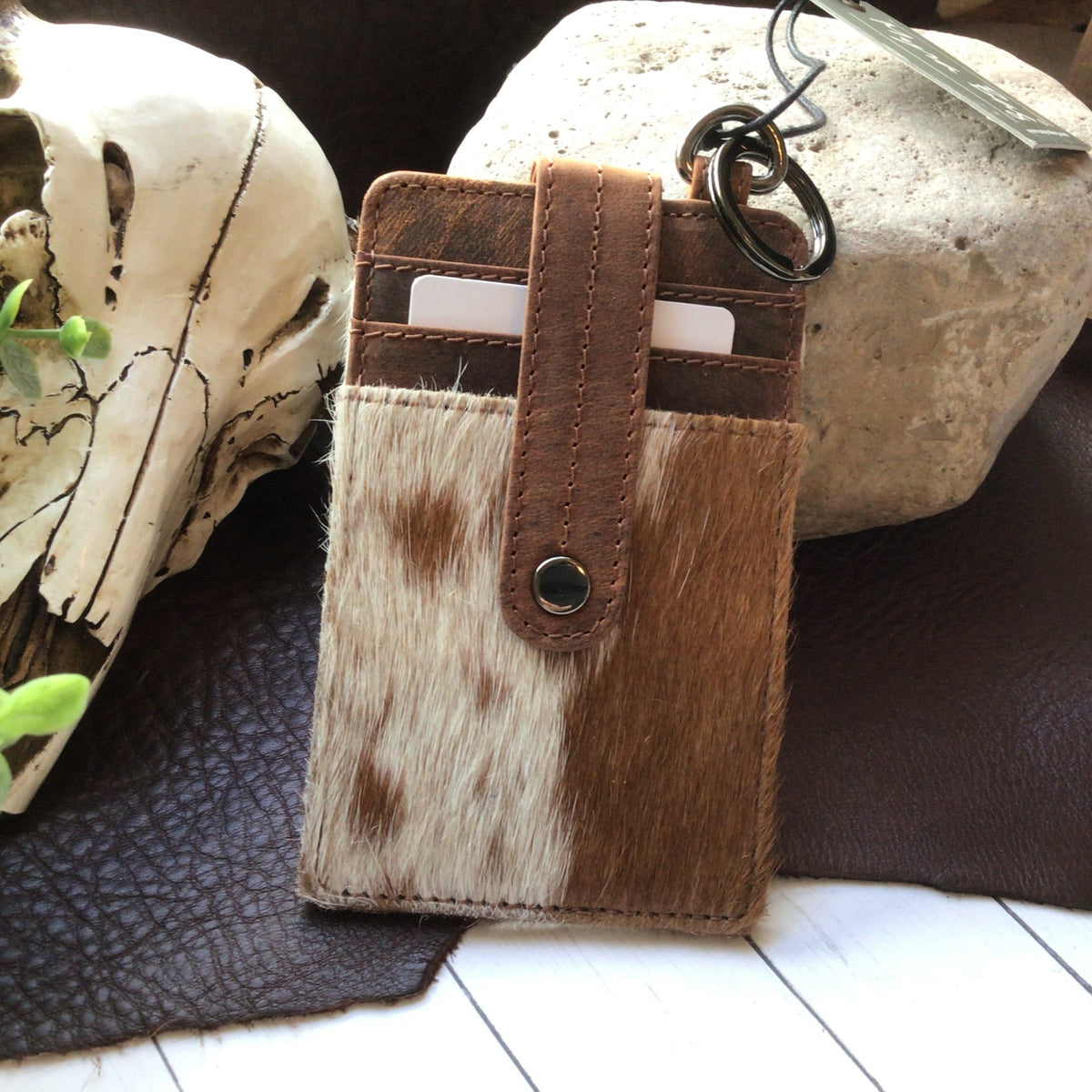 A small card holder wallet made of brown and cream cowhide with a brown leather strap and snap closure, shown with metal key rings. The wallet is styled on a dark leather surface with a skull, greenery, and a stone in the background.