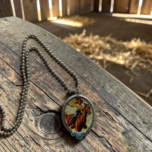 A silver-tone ball chain necklace with an oval pendant featuring a colorful illustration of a cowboy riding a horse, resting on a rustic wooden surface with a blurred barn interior in the background.