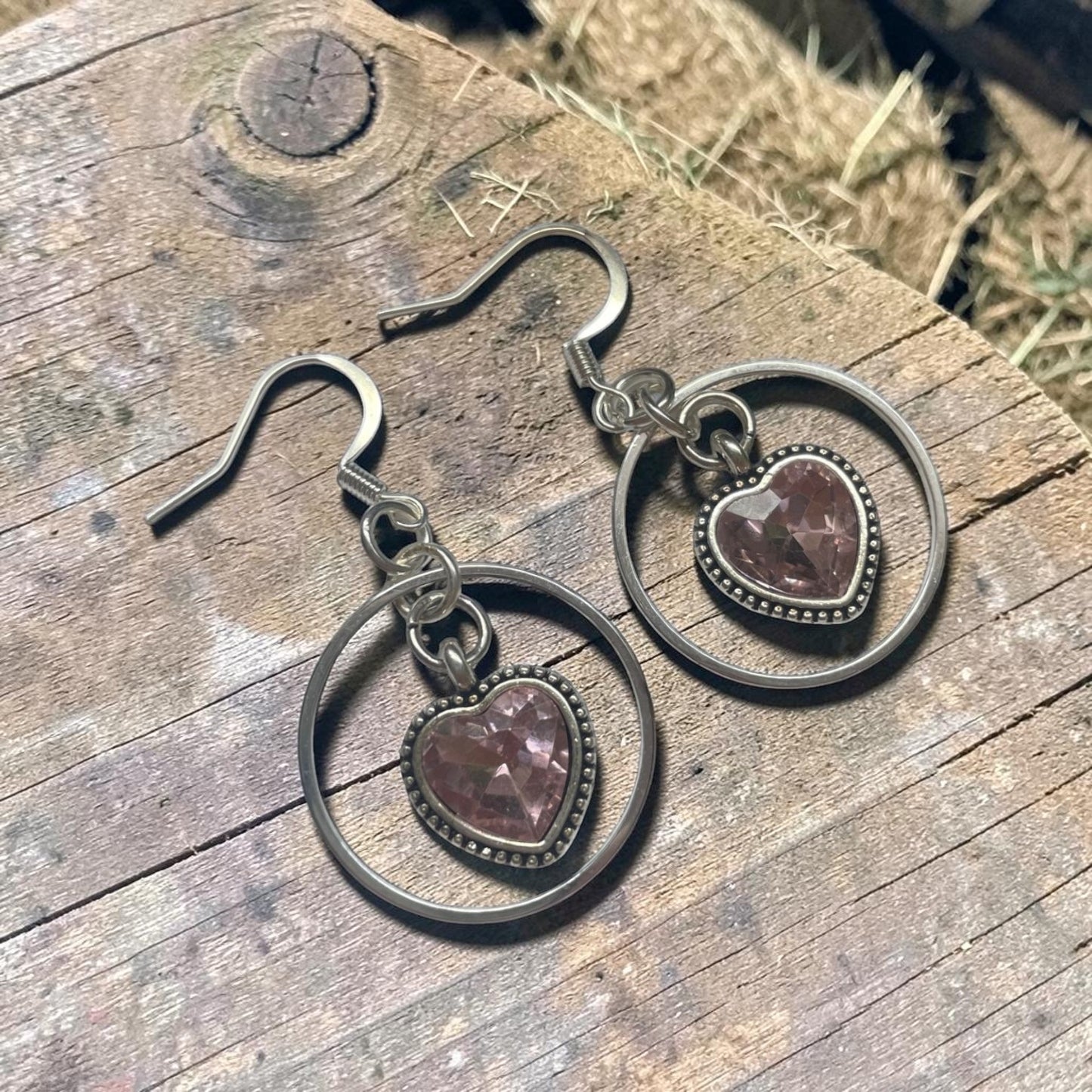 A pair of silver-toned dangle earrings with heart-shaped pinkish-purple stones, displayed on a rustic wooden surface.