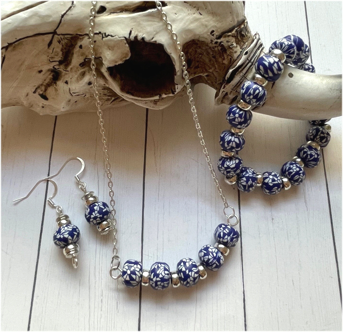 A jewelry set featuring a silver chain necklace, matching bracelet, and drop earrings, all adorned with blue and white patterned beads and small silver spacer beads, displayed on a white wood background with a piece of driftwood.