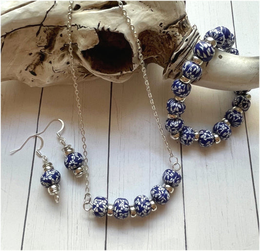 A jewelry set featuring a silver chain necklace, matching bracelet, and drop earrings, all adorned with blue and white patterned beads and small silver spacer beads, displayed on a white wood background with a piece of driftwood.