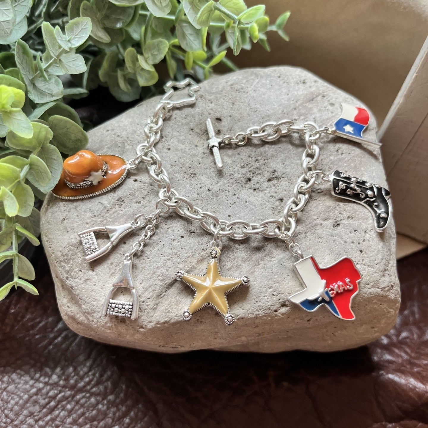 A silver charm bracelet displayed on a gray stone, featuring western-themed charms including a gold star, Texas flag, cowboy boots, and a cowboy hat, with green leaves and a brown leather surface in the background.