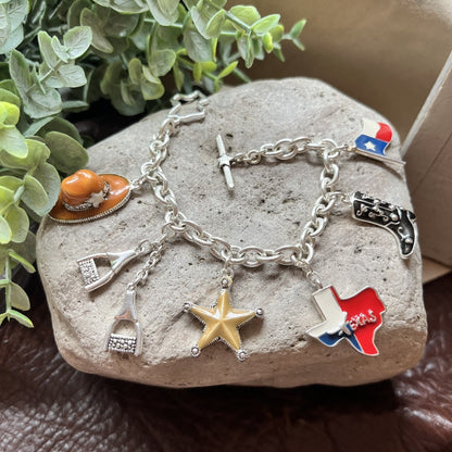 A silver charm bracelet displayed on a stone, featuring various western-themed charms including a cowboy hat, Texas state, boot, and star.