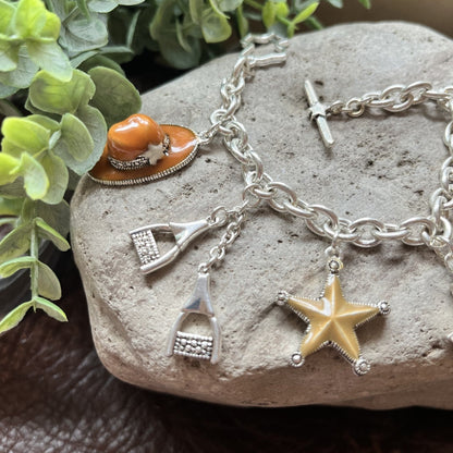 A silver-tone charm bracelet displayed on a textured stone, featuring a starfish charm, a small knife charm, a small hat charm, and a small rectangular charm, styled with green leaves in the background.