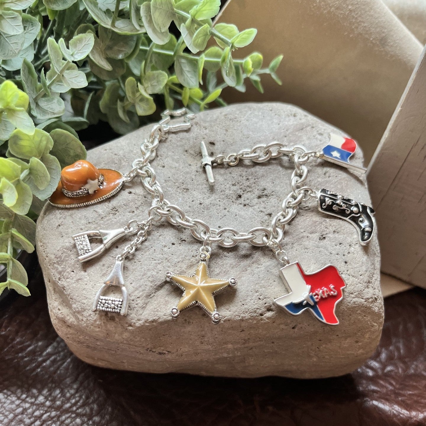 A silver charm bracelet displayed on a textured stone, featuring charms for a cowboy hat, horseshoe, lasso, star, cross, and Texas flag, with a small boot charm, set against a brown leather surface with green foliage in the background.