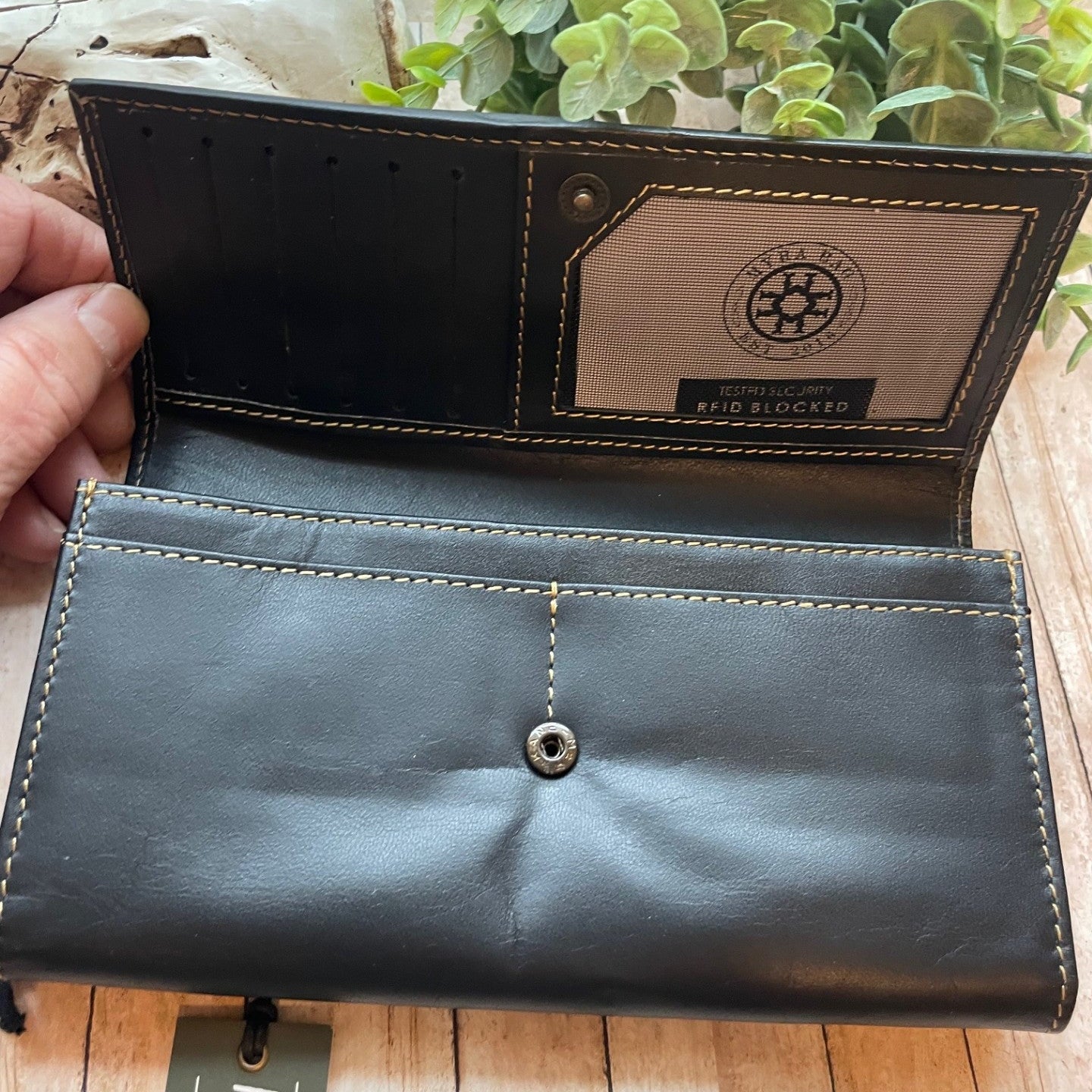 Black leather wallet with brand tag held open on a wooden surface with plants in the background