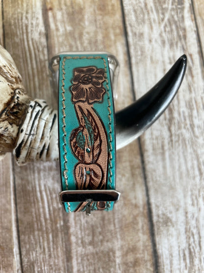 A turquoise leather watch band with hand-tooled brown floral and leaf designs, displayed on a rustic wood background with a horn and bone prop.