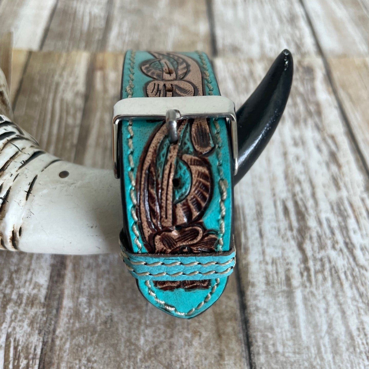 A turquoise leather bracelet with brown floral tooling and a silver buckle, displayed on a rustic wooden surface with a horn prop.