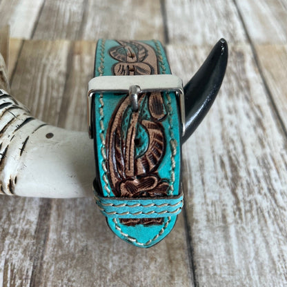 A turquoise leather bracelet with brown floral tooling and a silver buckle, displayed on a rustic wooden surface with a horn prop.